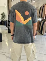 Remera oversize color negro con efecto gastado, manga corta y estampa artística en la espalda con diseño de montañas y sol en tonos amarillos, naranjas y verdes.