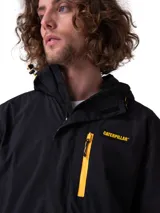 Chaqueta impermeable negra Caterpillar de tejido ripstop, con capucha ajustable, cierre frontal con cremallera y velcro, y bolsillos con cierre, destacando un bolsillo en el pecho con cremallera amarilla y parche con el logo de la marca.
