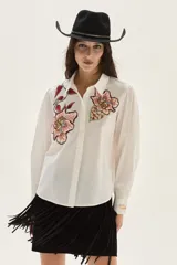 Camisa blanca de algodón con bordados florales en tonos rosados y rojizos en el frente. Presenta mangas largas abullonadas y puños con bordado.