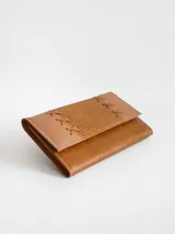 Billetera de cuero color camel con solapa frontal decorada con costuras en forma de cruz y textura grabada. Cuenta con cierre de broche y diseño rectangular.