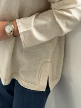 Blusa de lino color beige con escote en V, mangas largas con sutiles frunces en los hombros y costuras decorativas visibles en el cuello y el ruedo.
