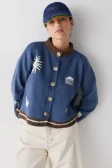 Cardigan de punto color azul con detalles bordados en blanco, cuello redondo y puños en color marrón. Presenta cierre frontal con botones grandes y un borde inferior con rayas horizontales en marrón y blanco.
