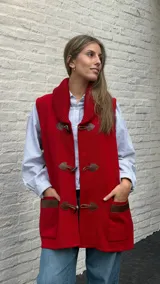 Chaleco largo de lana color rojo con cuello tipo chal acanalado. Presenta cierre frontal mediante tres trabas de cuero con botones tipo cuerno y bolsillos frontales con detalles en cuero.