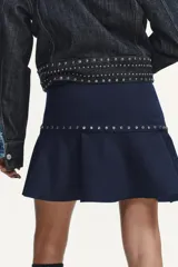Falda corta azul marino de tiro medio, con detalle de tachas plateadas y bajo con volante asimétrico. Cierre lateral con cremallera oculta.
