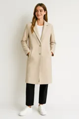 Tapado de paño color beige, de corte recto y largo hasta la rodilla. Presenta solapas clásicas, cierre frontal con dos botones y bolsillos laterales.