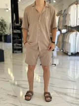 Camisa de manga corta color blanco marfil, confeccionada en tejido texturizado con cuello camisero, cierre frontal de botones y un pequeño bordado oscuro en el pecho.