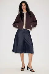 Chaqueta estilo bomber de color marrón oscuro, con cuello redondo, manga larga y cierre frontal con cremallera. Presenta bolsillos delanteros de vivo y un diseño con bajo fruncido.