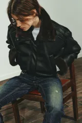 Cazadora bomber de efecto piel en color negro, con cuello subido, manga larga y bajo elástico. Presenta interior y puños con aplicación de pelo sintético en color marrón a contraste, cierre frontal con cremallera y botones a presión, y bolsillos laterales de vivo.