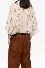 Camisa oversize de tejido fantasía color crema con estampado de pájaros y flores en tonos azules, amarillos, naranjas y rojos. Presenta finas rayas verticales en colores pastel y lentejuelas distribuidas por toda la prenda. Tiene cuello camisero, cierre frontal con botones y mangas cortas rectas con un ligero vuelo. Incluye un bolsillo de plastrón en el pecho izquierdo.