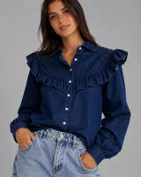 Camisa de denim azul oscuro con cuello camisero, cierre frontal de botones y detalle de volados en los hombros y el pecho. Presenta mangas largas con puños ajustados.