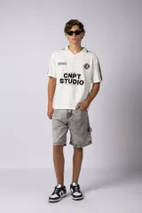 Remera tipo chomba de tejido de punto color blanco, con cuello en V y mangas cortas. Presenta rayas verticales sutiles en la trama y estampado gráfico frontal con la inscripción CNPT STUDIO y detalles en negro.