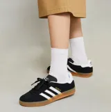 Championes Adidas Gazelle Indoor de diseño clásico, confeccionados en gamuza negra con las icónicas tres tiras laterales en blanco. Presentan el nombre del modelo en letras doradas, talón en contraste blanco y suela de caucho color caramelo.
