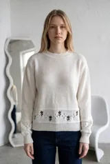 Sweater de punto color crema con cuello redondo, manga larga y diseño de calados en los puños. Presenta un detalle de bordado floral en la parte inferior y una franja con perforaciones decorativas.