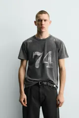 Camiseta de corte slim fit, cuello redondo y manga corta, color gris oscuro con efecto lavado. Presenta un gran estampado frontal con el número "74" en blanco desgastado y pequeños estampados circulares en los hombros.