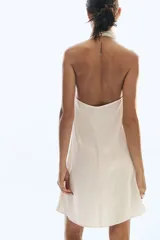 Minivestido de corte holgado en tejido fluido de satén color beige, con escote drapeado y tira halter con cierre de botones.