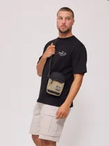 Bandolera negra de poliéster con logo de la marca Rusty en relieve en el frente. Cuenta con un bolsillo frontal con solapa y cierre de cremallera en el compartimento principal. La correa es ajustable.