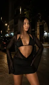 Vestido corto de dos piezas compuesto por un top interno tipo strapless y un vestido de tul transparente con mangas largas acampanadas. Presenta un diseño ajustado con frunces que estilizan la figura y detalle metálico en el top.