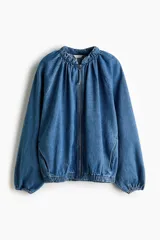 Campera bomber oversize de denim azul, con cuello elevado fruncido, cierre frontal y bolsillos laterales. Cuenta con mangas raglán y elásticos en puños y bajo.