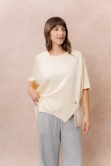 Buzo oversize color beige, con escote redondo, mangas cortas amplias de corte raglán y terminación frontal asimétrica en punta con aberturas laterales.