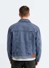 Chaqueta de jean estilo boxy, con corte amplio y cuadrado, solapas clásicas, cierre frontal de botones metálicos, bolsillos frontales con solapa y detalles desflecados en los bordes.
