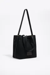 Bolso shopper de estructura blanda confeccionado en tejido de malla técnica negra. Cuenta con doble asa de hombro, cierre mediante cordón ajustable y etiqueta frontal con el logo de la marca Bimba y Lola.