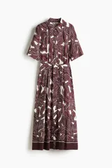 Vestido midi con cuello camisero, botonadura frontal y mangas cortas. Presenta un estampado abstracto en tonos marrón oscuro y blanco, elástico en la cintura y cinturón para atar.