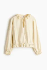 Blusa de viscosa color amarillo claro con detalles de broderie, cuello con volados y abertura frontal en V con tirantes finos. Presenta mangas globo largas y ribetes con volados en los puños y el dobladillo.
