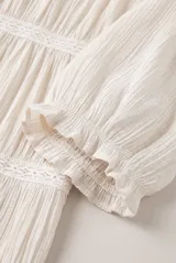 Camisa Belen de manga corta abullonada, color beige, confeccionada en tejido liviano con textura. Presenta cuello redondo con solapa y delicadas franjas horizontales de puntilla y encaje, con cierre frontal de botones.