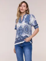 Blusa de crepe color blanco con estampado paisley en tonos azules y negros. Presenta cuello mao con escote en V, cierre frontal con botones y mangas cortas abullonadas con puños elastizados en punto smock.