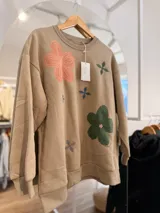 Buzo de cuello redondo en color beige, con diseño oversize y felpa interna. Presenta bordados de flores en tonos rosa, verde y azul distribuidos en el frente.