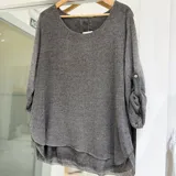 Sweater de punto color gris visón, con cuello redondo, mangas largas con trabilla para sujetar arremangadas y dobladillo asimétrico en capas de tela ligera.