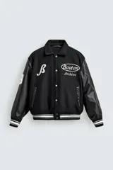 Cazadora estilo varsity con cuerpo de tejido con lana y mangas de efecto piel. Presenta cuello solapa, cierre frontal con botones a presión, bolsillos laterales y puños con elástico a rayas. Incluye parches y bordados decorativos en el frente.