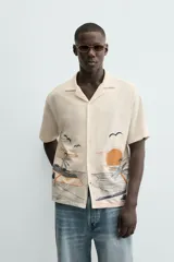Camisa de manga corta y calce relajado en color beige, con cuello bowling y cierre frontal de botones. Presenta un diseño bordado en la parte inferior con motivos de paisaje tropical que incluyen palmeras, sol y aves.