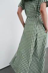 Vestido midi camisero de cuello solapa y escote pico, con estampado de cuadros vichy en verde oscuro y blanco. Presenta manga corta con vuelta, bolsillos laterales ocultos, solapas decorativas en el pecho, cinturón de lazo en el mismo tejido y cierre frontal con botones.
