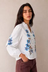 Camisa blanca de manga larga con bordados florales en azul, escote en V y cuello con volados fruncidos.