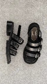 Sandalias de cuero negro con punta semicuadrada, taco cuadrado bajo y tres tiras frontales con hebillas metálicas plateadas. Cuentan con tira trasera con cierre ajustable.