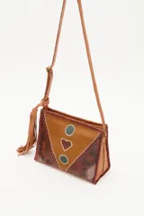 Cartera bandolera de cuero vacuno con diseño patchwork que combina texturas lisas y efecto reptil. Presenta detalles decorativos en forma de círculos y un corazón central, con costuras visibles en los bordes y una borla colgante en el lateral. Incluye correa larga ajustable.