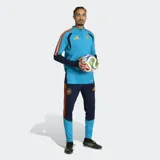 Buzo de entrenamiento Adidas Tiro España 26, color celeste con detalles en azul marino y vivos con los colores de la bandera española en los hombros. Presenta logo de Adidas en naranja y escudo de España bordado. Cuenta con cierre frontal parcial y tecnología Formotion para mayor libertad de movimiento.