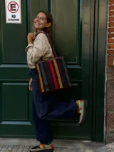 Cartera tipo tote de cuero con diseño de franjas verticales multicolores en tonos tierra, rojo y azul. Cuenta con correas trenzadas de cuero y cierre superior.