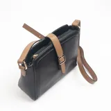 Bandolera de cuero sintético negro con textura granulada, cuenta con una solapa decorativa central en color tostado con hebilla metálica dorada y correa larga ajustable en combinación de colores negro y tostado.