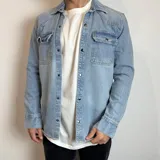 Chaqueta de jean en tono azul claro, con cierre frontal de botones metálicos a presión, dos bolsillos frontales con solapa y botones, y cuello clásico.