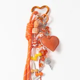 Llavero decorativo en tonos naranja, compuesto por una cuerda trenzada, un mosquetón en forma de corazón y múltiples dijes colgantes que incluyen cuentas de cristal, figuras de flores, un dije de corazón de cuero sintético y una esfera brillante.