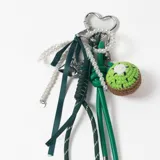 Llavero decorativo con mosquetón en forma de corazón, compuesto por una cadena de perlas, cintas de tela verde, cordones técnicos y un dije tejido a crochet con forma de kiwi.