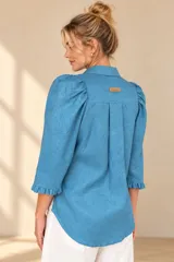 Camisa de lino color blanca, con cuello clásico, mangas tres cuartos abullonadas en los hombros y terminación con volados en los puños, cierre frontal con botones ocultos.
