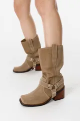 Bota de caña media confeccionada en cuero gamuzado color chocolate. Presenta un diseño de estilo western con taco cuadrado de 7 cm y una correa decorativa en el tobillo con herrajes metálicos plateados.