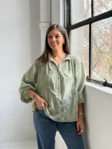 Camisa verde claro de corte oversize, con mangas tres cuartos abullonadas, escote en V con cuello camisero y cierre frontal parcial de botones. Cuenta con delicados bordados florales tono sobre tono distribuidos por toda la prenda.