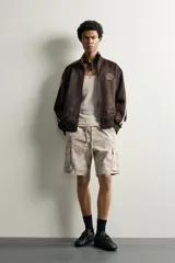 Cazadora relaxed fit de tejido efecto ante color marrón oscuro, con cuello subido y cierre frontal de cremallera. Presenta mangas largas con bandas blancas a contraste en los hombros, bolsillos laterales de vivo, y bordados en hilo blanco en el pecho.