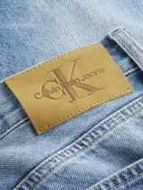 Jeans de mezclilla azul claro con efecto lavado y desgaste sutil. Presentan un corte recto y talle medio, con cierre de cremallera y diseño de cinco bolsillos. Incluyen una etiqueta de cuero con el logo de Calvin Klein en el bolsillo trasero derecho.