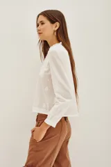 Camisa blanca de algodón con cuello alto, cierre frontal de botones ocultos y detalles de bordado floral en el pecho. Presenta mangas largas con puños elásticos y volados decorativos en la parte frontal.