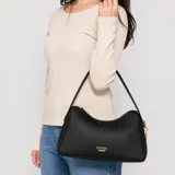 Cartera Guess modelo Jasmin de color negro, confeccionada en símil cuero con textura granulada. Presenta diseño estructurado con cierre superior y herrajes metálicos en tono dorado, incluyendo el logo de la marca en la parte frontal y una manija única para llevar al hombro.